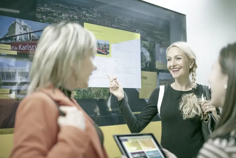 Eine Frau zeigt zwei Personen etwas auf einem großen Display. Bild: KTG Karlsruhe Tourismus GmbH